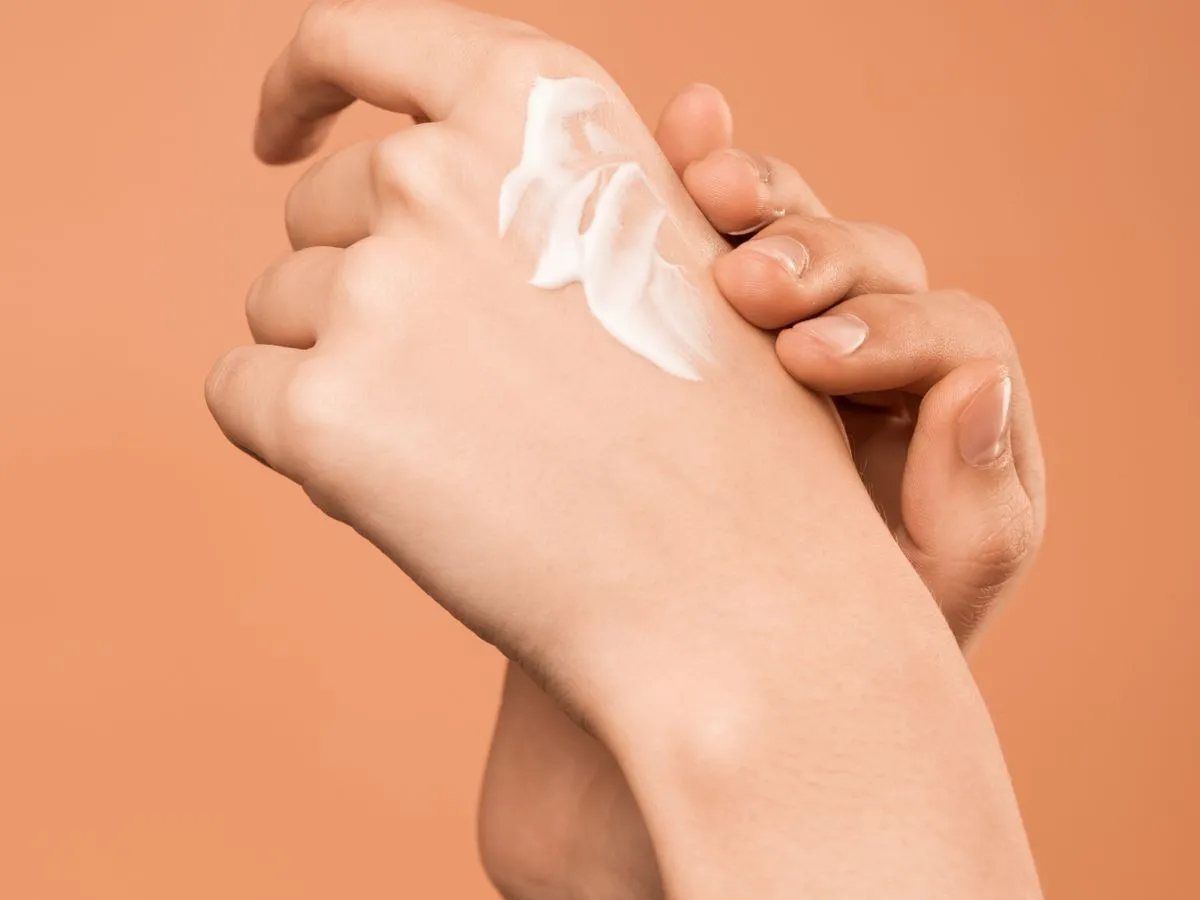 Manos con una porción de crema hidratante extendida sobre el dorso de la mano, sobre fondo color terracota